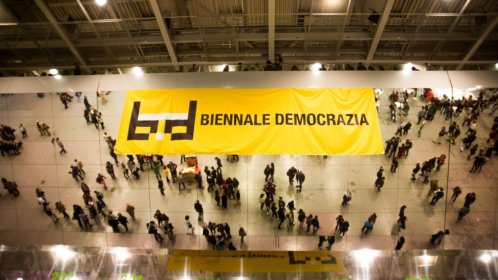 Il logo di Biennale Democrazia su uno striscione appeso sopra a uno specchio che riflette il pubblico