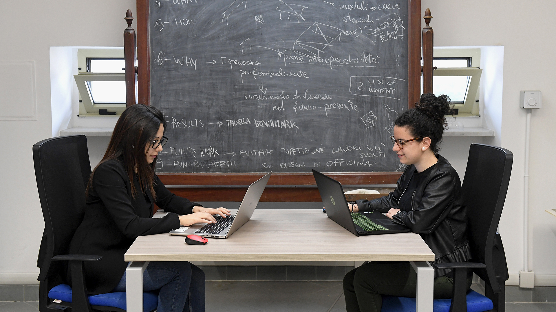 Due ragazze lavorano al computer davanti a una lavagna con formule scritte